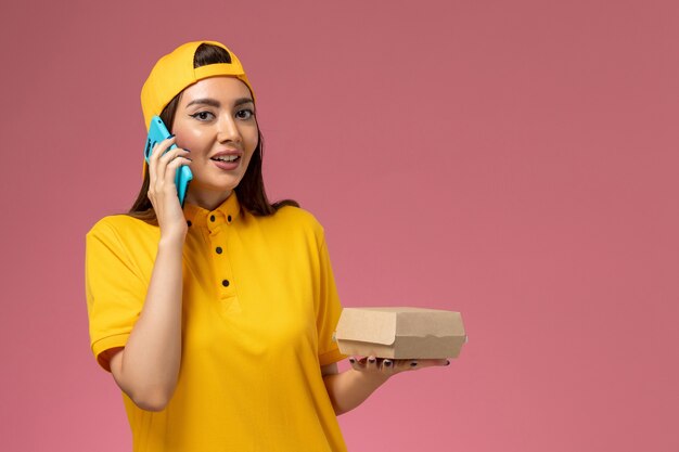 Вид спереди женщина-курьер в желтой форме и накидке, держащая пакет с едой и разговаривающая по телефону на светло-розовой стене, девушка службы доставки униформы компании
