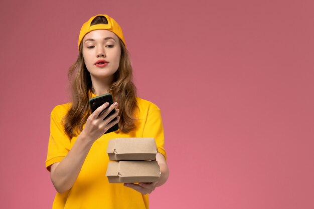 Вид спереди женщина-курьер в желтой униформе и накидке держит пакеты с продуктами, разговаривает по телефону на розовой стене.