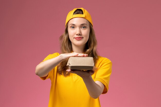 Front view female courier in yellow uniform and cape holding little delivery food package on light pink desk service delivery uniform girl job