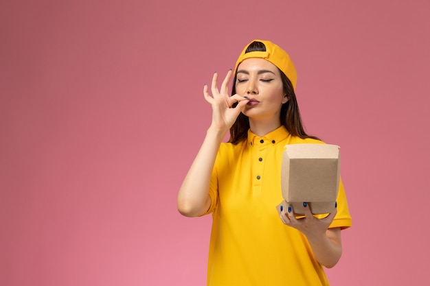 Front view female courier in yellow uniform and cape holding little delivery food package on light-pink wall service uniform delivery company girl job