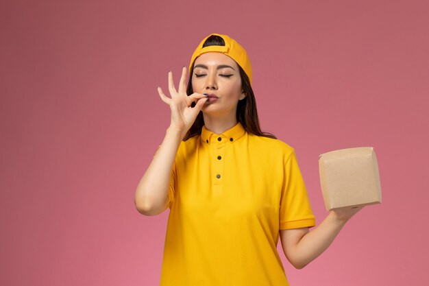 黄色の制服とケープの正面図の女性の宅配便は、淡いピンクの壁の制服サービス配達会社の女の子に小さな配達食品パッケージを保持しています