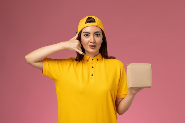 Вид спереди женщина-курьер в желтой униформе и плаще, держащая небольшой пакет с доставкой еды на светло-розовой стене, униформа, служба доставки, работа, работа