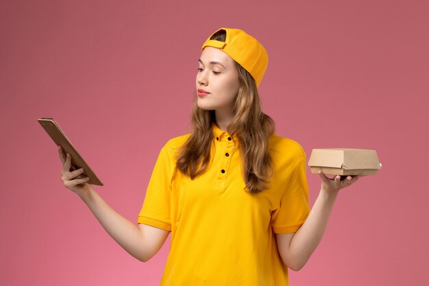 Front view female courier in yellow uniform and cape holding little delivery food package and notepad on light pink wall service delivery uniform work