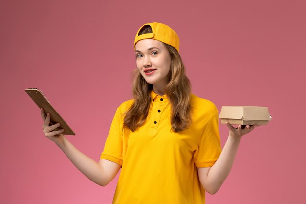 Front view female courier in yellow uniform and cape holding little delivery food package and notepad on light pink wall service delivery uniform worker