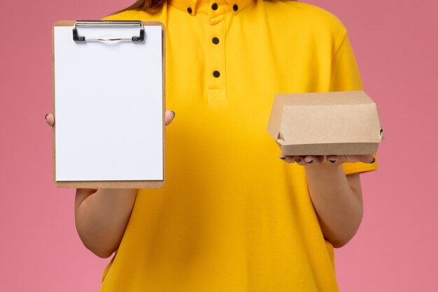 Corriere femminile di vista frontale in uniforme gialla e mantello che tiene piccolo pacchetto di cibo per la consegna e blocco note sul lavoro di consegna uniforme di servizio a parete rosa chiaro