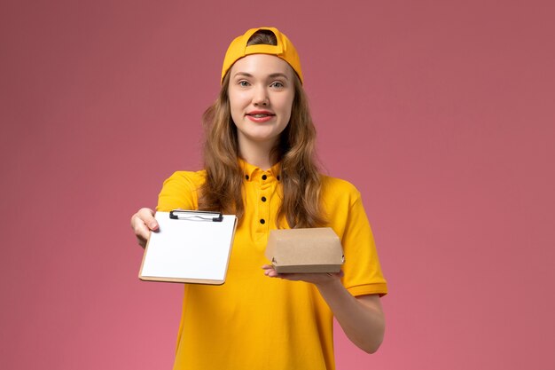 黄色の制服とピンクの壁に小さな配達食品パッケージのメモ帳を保持している岬の正面図の女性の宅配便仕事仕事配達制服