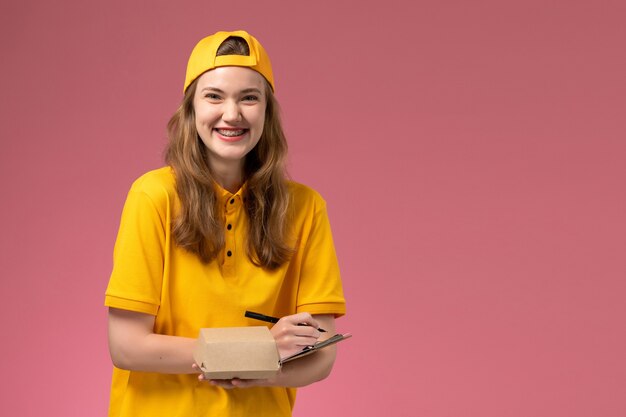 Front view female courier in yellow uniform and cape holding little delivery food package and notepad writing on light pink wall service delivery uniform