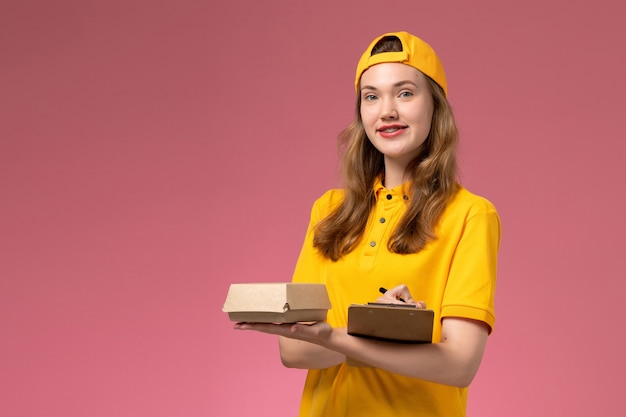 Вид спереди женщина-курьер в желтой униформе и накидке с маленькой ручкой для доставки еды и блокнотом на светло-розовой стене работник службы доставки