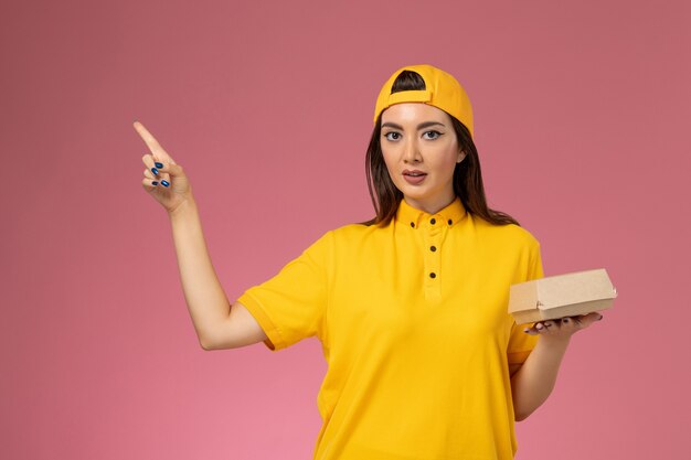 Front view female courier in yellow uniform and cape holding little delivery food package on pink wall uniform girl service delivery company