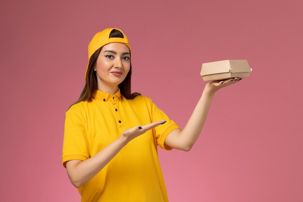 ピンクの壁の制服サービス配達会社の女の子に小さな配達食品パッケージを保持している黄色の制服と岬の正面図の女性の宅配便