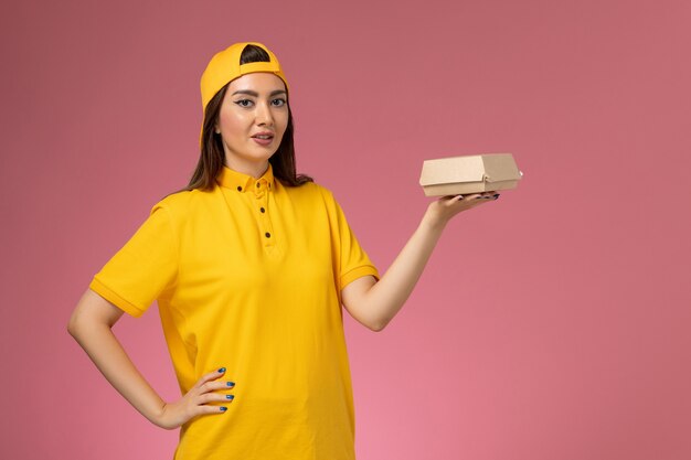 Front view female courier in yellow uniform and cape holding little delivery food package on the pink wall uniform service delivery company job work