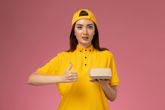 Front view female courier in yellow uniform and cape holding little delivery food package on the pink wall uniform service delivery company job worker work