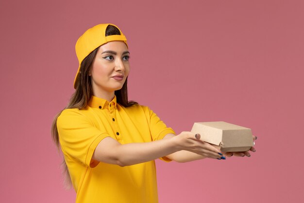 Front view female courier in yellow uniform and cape holding little delivery food package on pink wall uniform service delivery company job worker