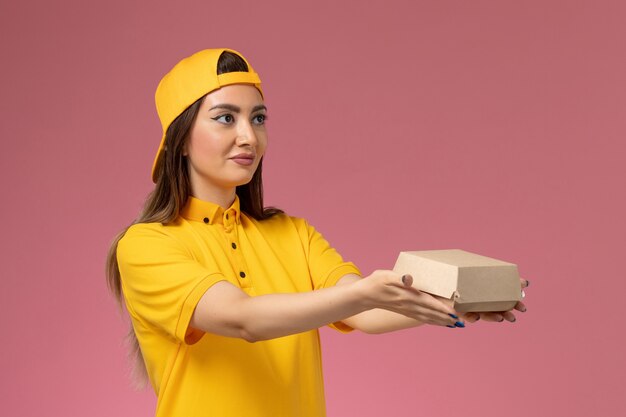 ピンクの壁の制服サービス配達会社の仕事の労働者に小さな配達食品パッケージを保持している黄色の制服と岬の正面図の女性の宅配便