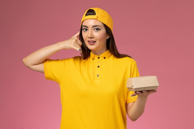 Вид спереди женщина-курьер в желтой униформе и плаще с маленьким пакетом еды для доставки на розовой стене.