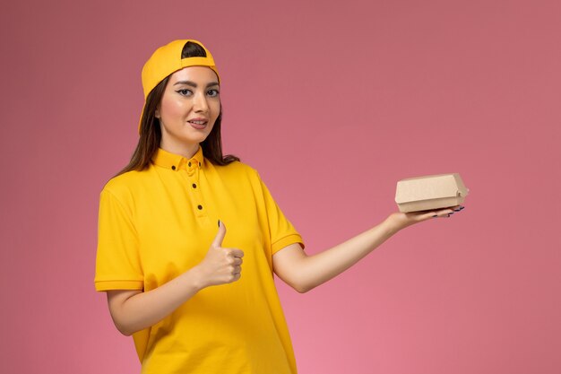 분홍색 벽 유니폼 서비스 배달 작업 작업자 작업에 작은 배달 음식 패키지를 들고 노란색 유니폼과 케이프의 전면보기 여성 택배