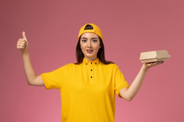 Front view female courier in yellow uniform and cape holding little delivery food package on the pink wall uniform service delivery worker