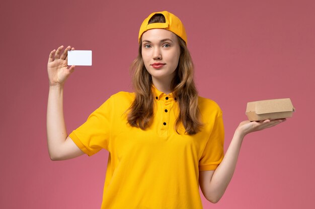 ピンクの壁のサービス配達労働者の制服に小さな配達食品パッケージとプラスチックカードを保持している黄色の制服と岬の正面図の女性の宅配便