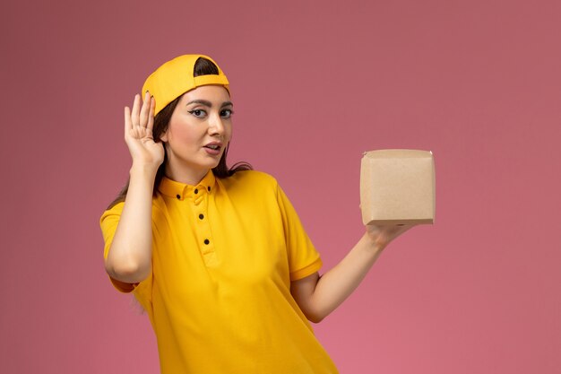 Front view female courier in yellow uniform and cape holding little delivery food package trying to hear on light-pink wall uniform service delivery company