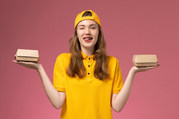 Front view female courier in yellow uniform and cape holding little delivery food packages on light pink wall service job delivery uniform