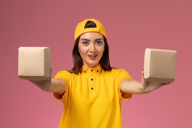 Front view female courier in yellow uniform and cape holding little delivery food packages on the light-pink wall service uniform delivery