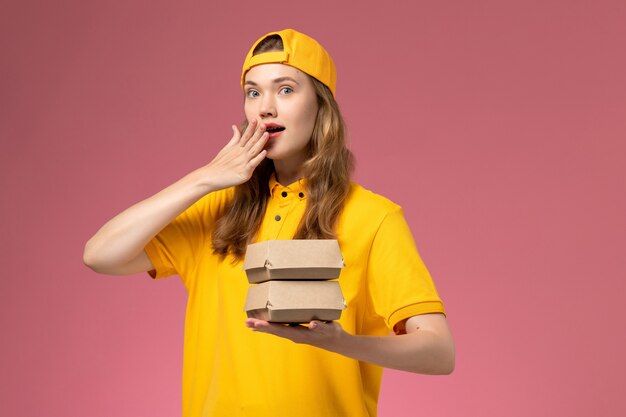 Front view female courier in yellow uniform and cape holding little delivery food packages on the pink wall service worker delivery uniform