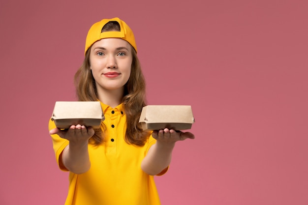 Front view female courier in yellow uniform and cape holding little delivery food packages smiling on the pink wall service worker delivery uniform