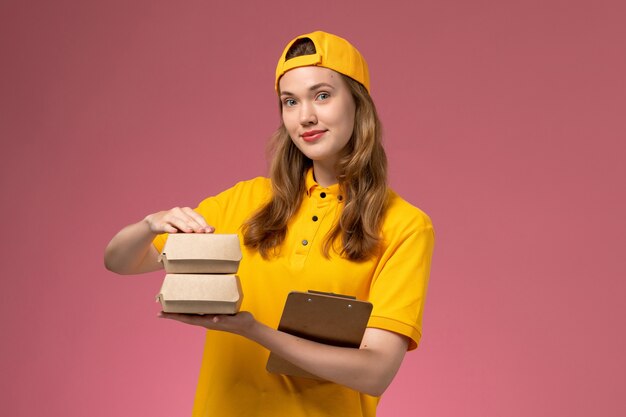 Front view female courier in yellow uniform and cape holding little delivery food packages with notepad on pink wall service delivery uniform work