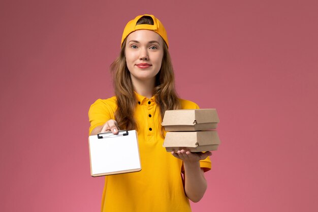 Front view female courier in yellow uniform and cape holding little delivery food packages with notepad on pink wall service delivery uniform worker