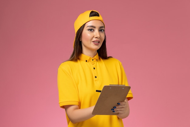 Front view female courier in yellow uniform and cape holding notepad and writing on light pink wall company service uniform delivery worker