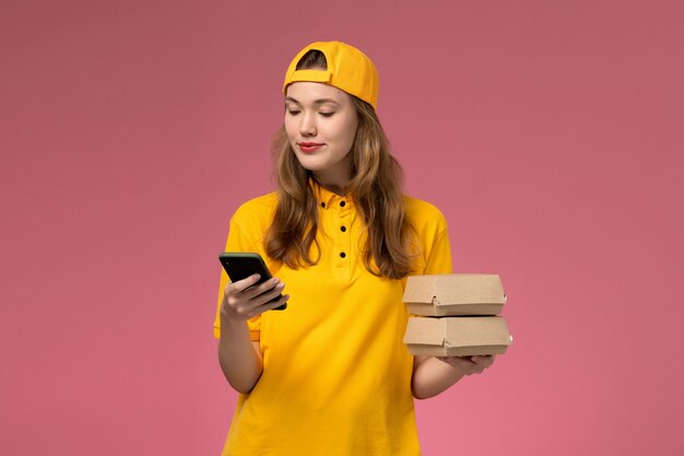 노란색 유니폼과 케이프 핑크 벽 회사 서비스 배달 유니폼 작업에 음식 패키지와 전화를 들고 전면보기 여성 택배