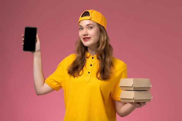 노란색 유니폼과 케이프 핑크 벽 회사 서비스 배달 유니폼에 음식 패키지와 전화를 들고 전면보기 여성 택배