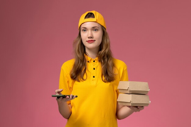 黄色の制服を着た正面図の女性の宅配便とピンクの壁の会社のサービスワーカーの配達制服に食品パッケージと電話を保持している岬