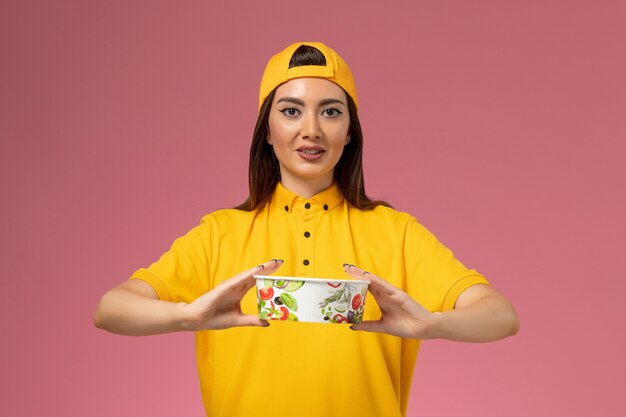 Front view female courier in yellow uniform and cape holding round delivery bowl on pink wall service worker uniform delivery