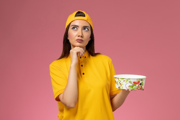 Front view female courier in yellow uniform and cape holding round delivery bowl thinking on pink wall service uniform delivery