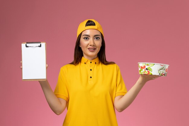 Front view female courier in yellow uniform and cape holding round delivery bowl with notepad on pink wall service uniform delivery job