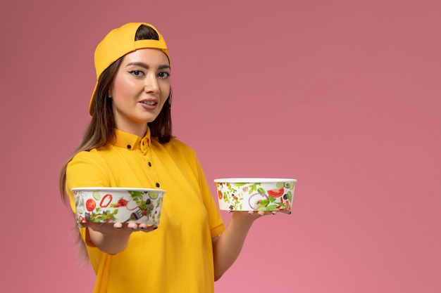 Free photo front view female courier in yellow uniform and cape holding round delivery bowls on pink wall service uniform delivery job work