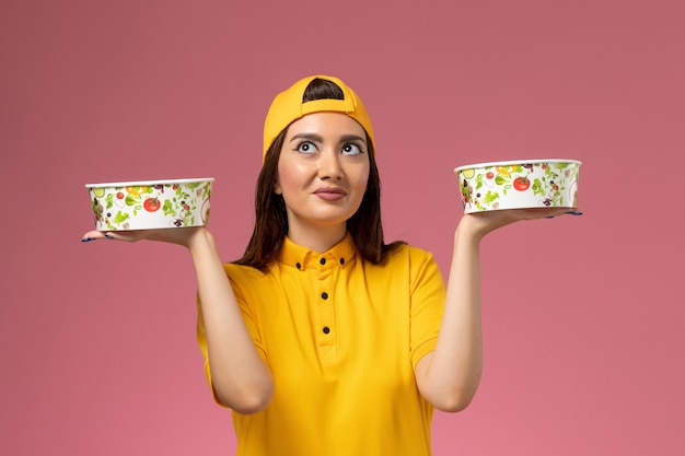 Front view female courier in yellow uniform and cape holding round delivery bowls on pink wall service uniform delivery job
