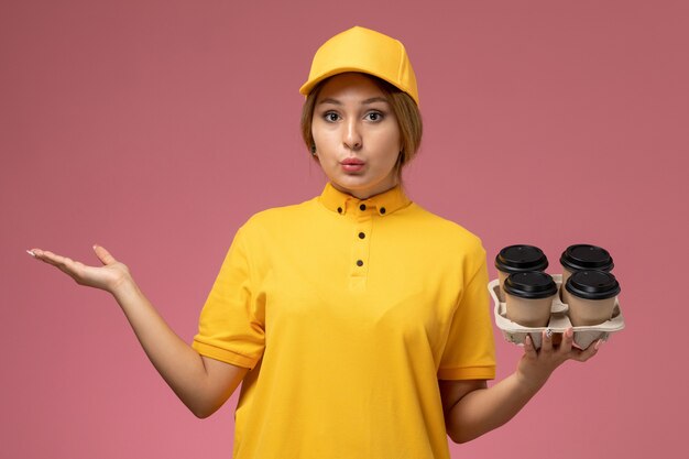 ピンクの背景に茶色のプラスチック製のコーヒーカップを保持している黄色の制服黄色のケープの正面図女性宅配便制服配達作業色の仕事