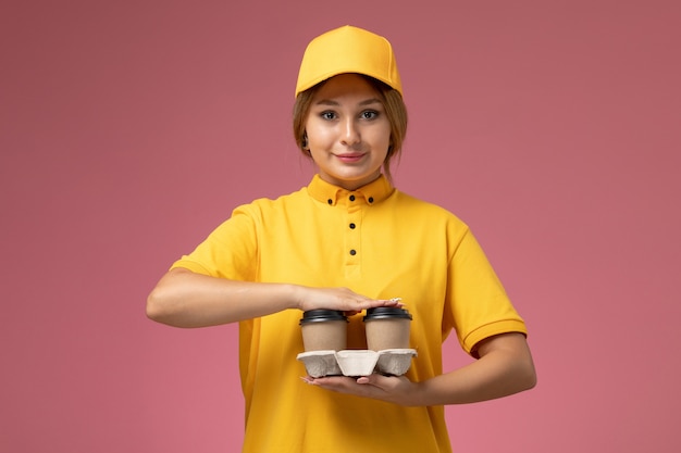 Вид спереди женщина-курьер в желтой форме с желтым плащом держит коричневые пластиковые кофейные чашки на розовом фоне.