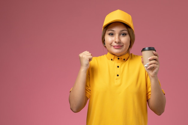 Front view female courier in yellow uniform yellow cape holding coffee cup on the pink desk uniform delivery female