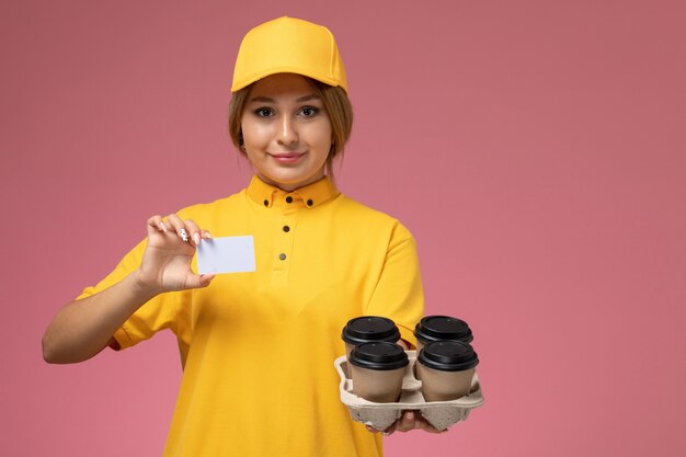 Вид спереди женщина-курьер в желтой форме с желтым плащом держит кофейные чашки и карточку на розовом фоне.
