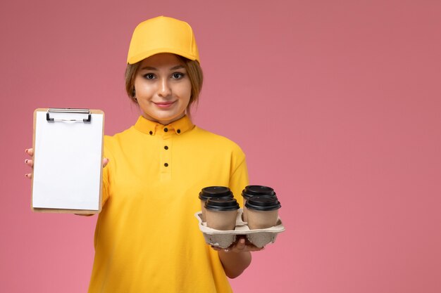 Front view female courier in yellow uniform yellow cape holding coffee cups notepad on pink background uniform delivery work color job