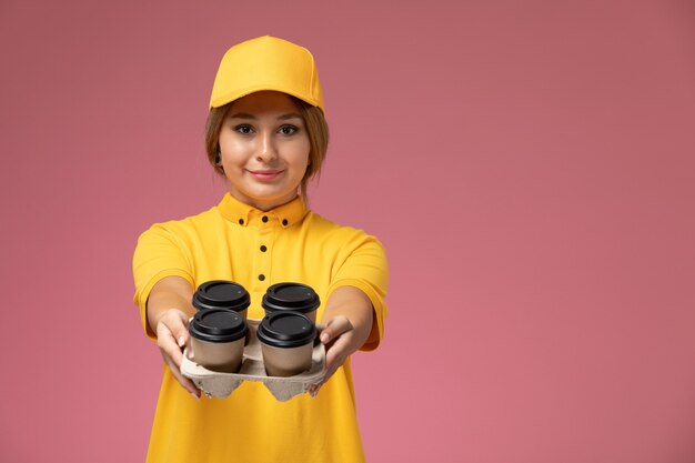 Вид спереди женщина-курьер в желтой униформе, желтой накидке с кофейными чашками на розовом фоне.