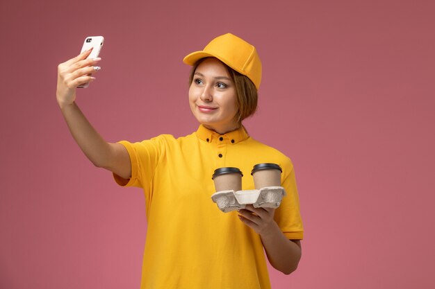 Front view female courier in yellow uniform yellow cape holding coffee cups and taking a selfie on pink background uniform delivery work color job
