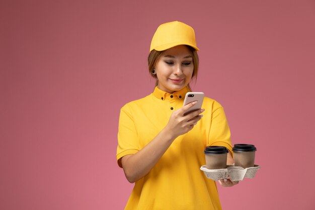 ピンクの背景に電話を使用してコーヒーカップを保持している黄色のユニフォーム黄色のケープの正面図女性宅配便