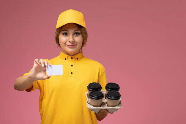 분홍색 배경에 균일 한 배달 작업 색상에 커피 컵과 흰색 카드를 들고 노란색 유니폼 노란색 케이프에서 전면보기 여성 택배