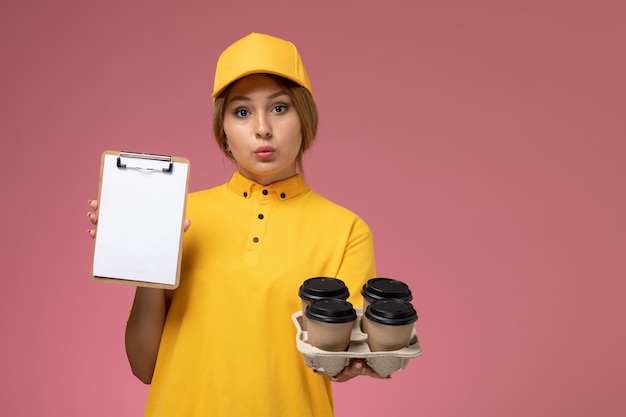 ピンクの背景にコーヒーカップと白いメモ帳を保持している黄色の制服黄色のケープの正面図女性宅配便制服配達作業色の仕事