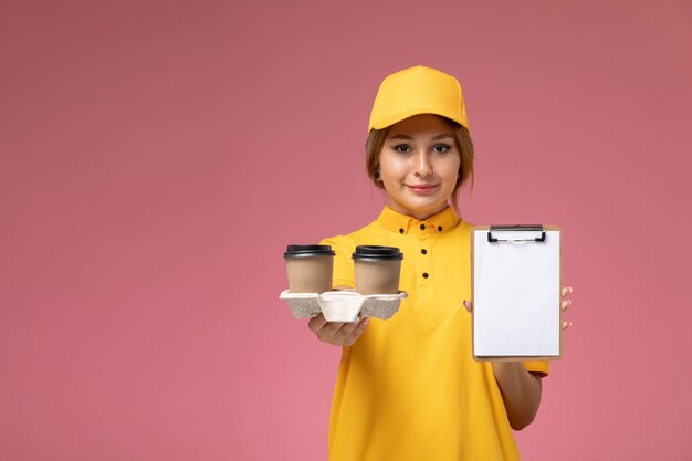 Free photo front view female courier in yellow uniform yellow cape holding coffee and notepad on pink background uniform delivery job