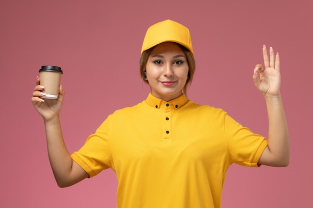 ピンクの背景の制服配達の仕事で笑顔でコーヒーを保持している黄色の制服黄色の岬の正面図女性宅配便